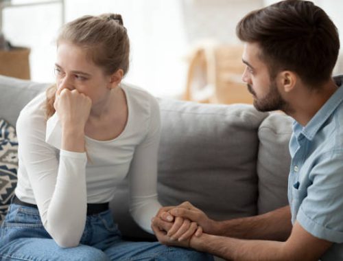 Loving young husband hold crying wife hand showing empathy and support, millennial couple sit on couch at home reconcile after fight, caring man making peace with beloved woman. Relationships concept